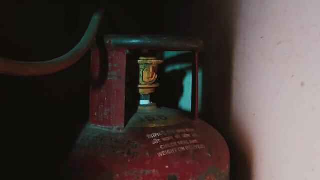 An orange LPG cylinder, essential for Indian kitchens, rests in a dim alcove. Its yellow regulator is set for cooking, despite the 2026 supply crisis and rising costs making home meals a challenge.