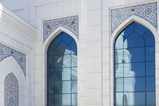 White mosque facade with pointed arch windows and floral tilework at Minor Mosque in Tashkent. Reflections of a minaret visible in blue glass panels, ideal for travel and architectural publications.