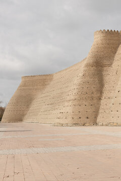 Ark fortress walls in Bukhara with massive sloping mud-brick ramparts and crenellated battlements under an overcast sky. Ideal for travel guides, history publications, and Central Asian tourism.