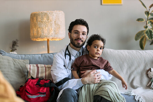 Portrait of young adult male doctor holding toddler during home visit consultation, looking at camera