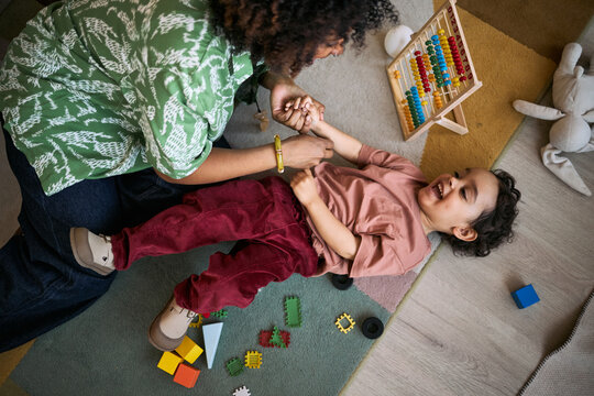Overhead shot of Black mother playing with toddler on floor, holding hands and laughing during home learning time