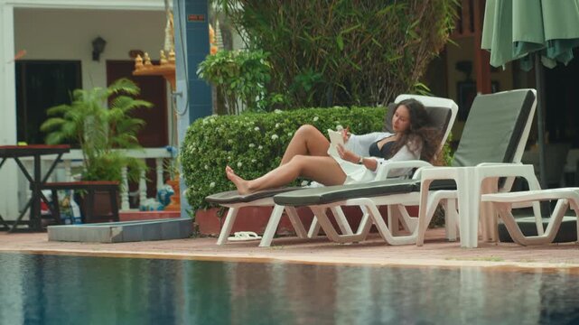 Woman enjoys tranquil island vacation. Serene woman reading by tropical pool. Peaceful woman unwinding with book beside reflective water. Woman leisurely resting near pool on tropical getaway day