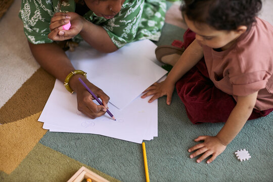 High angle view of Black mother guiding toddler drawing on paper during floor play, supporting early learning and bonding