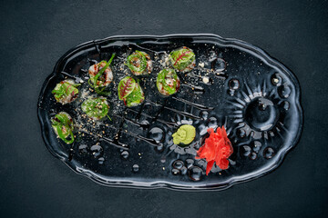 Sushi maki rolls topped with seaweed salad (chuka), sesame seeds, and balsamic glaze, served with ginger and wasabi on a dark plate © ismishko