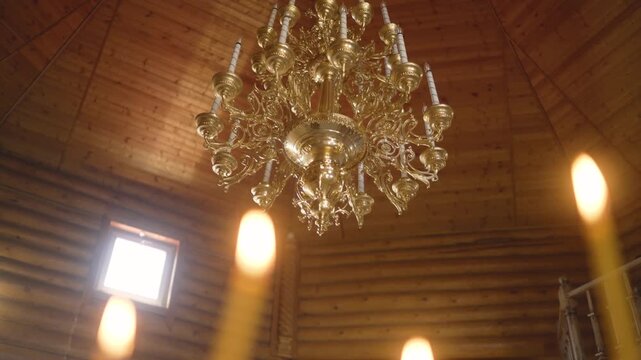 Low angle shot of large golden chandelier hanging from wooden ceiling in church. Warm light and blurred candles in foreground create peaceful, reverent atmosphere. Slow camera movement
