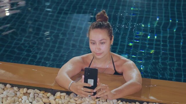 Poolside woman checking smartphone messages, relaxed expression leaning on pool edge, mosaic blue tiles and pebble border, nighttime glow and soft lights, casual swimsuit, scrolling social feed