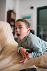 Fototapeta premium Young child lying on a couch with an open mouth, looking up in surprise or wonder while watching content on a screen in a domestic living room interior