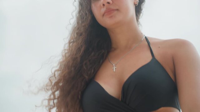 Fashionable woman on beach enjoying sun. Stylish darkskinned lady confidently posing on sunny coastline. Beautiful woman with windtousled hair dressed in swimwear by sunny island shore