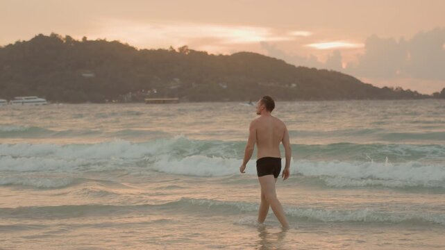 Man walking along island beach at golden hour, warm glow over rolling waves, active shoreline stroll, casual trunks and footsteps in wet sand, distant hills and breeze, leisure exploration