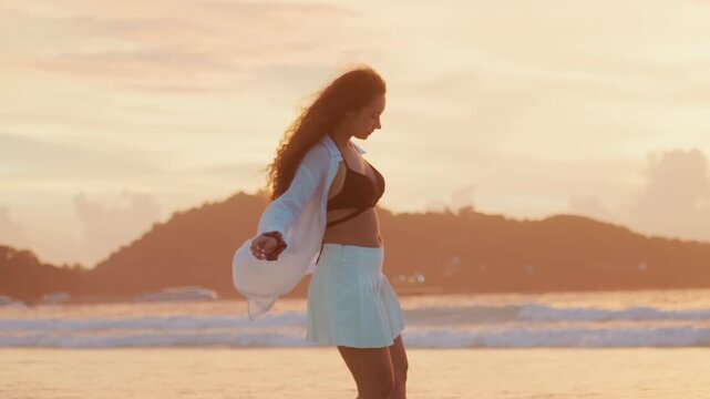 Black woman walking island beach sunset, model strolling along shoreline wearing bikini top, skirt and light coverup, windswept hair catching golden light, distant island silhouette over calm ocean,