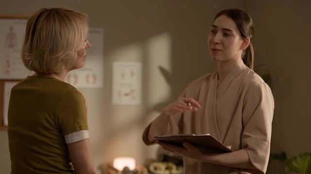 Side view medium shot of woman explaining neck and shoulder discomfort to massage therapist before session while therapist listening and taking notes on clipboard in cozy treatment room