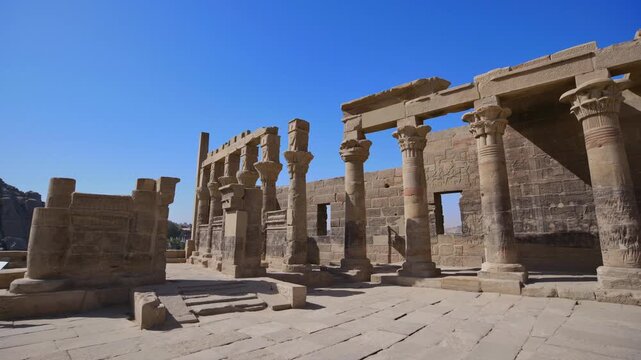 Ancient Philae Temple with towering walls covered in hieroglyphs and carved Hathor faces, captured under bright sunlight and clear blue sky.
