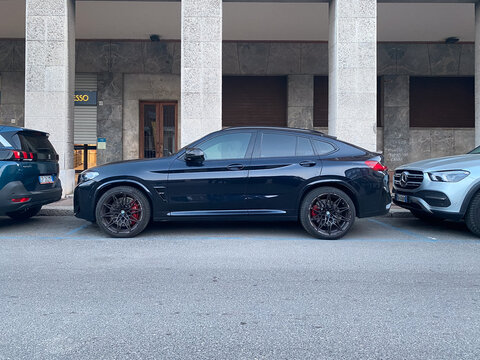 BMW X4 M Sport black luxury SUV parked on a European city street between Peugeot and Mercedes Benz, sleek coupe SUV profile, urban setting