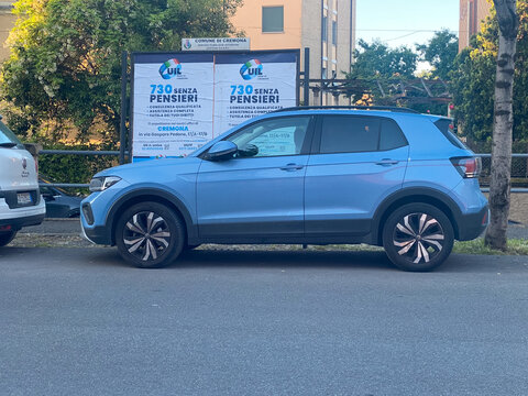 Light blue Volkswagen T Cross compact SUV parked on a Cremona city street beside a UIL 730 senza pensieri billboard, daytime