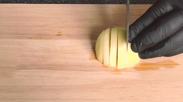 Chef in black gloves slicing fresh onion on wooden cutting board, cooking process close up.