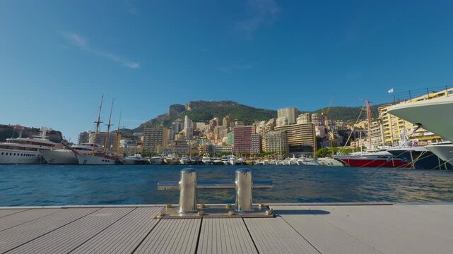 Static shot of Port Hercule in Monaco with free mooring cleat in foreground, luxury yachts, calm blue water, and La Condamine skyline against mountains, highlighting stillness and upscale setting.