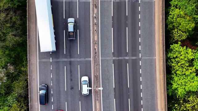Straight down aerial drone shot of white HGV articulated lorry changing lanes on M6 motorway, UK. Red truck and cars visible on multi-lane highway between green trees.