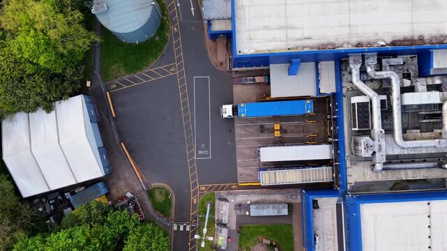 Straight down aerial drone shot of articulated HGV truck reversing into warehouse loading dock, UK. Blue and white lorries at distribution centre with yellow markings.