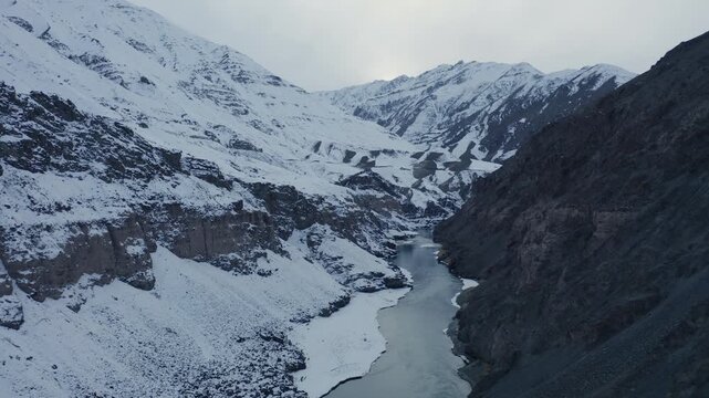 Indus River winds through a snow-covered mountain valley near Hemis National Park during winter. Slow forward aerial approach reveals the glacial river between steep Himalayan slopes.