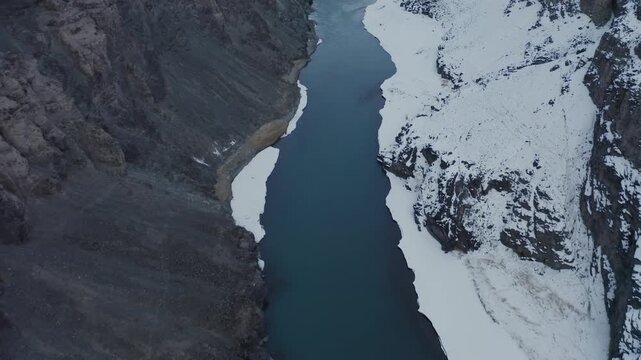 Indus River cuts through snow-covered terrain in Leh Valley, Ladakh, during winter season. Overhead bird's eye aerial view captures the contrast between dark mountain, water and white ice banks.