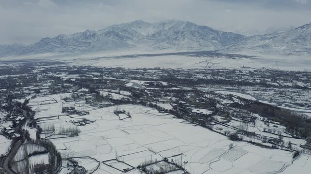 Indus River valley in Leh, Ladakh, India lie beneath snow-covered mountains during winter. The aerial view reveals agricultural fields blanketed in snow with Matho Village and the distance.