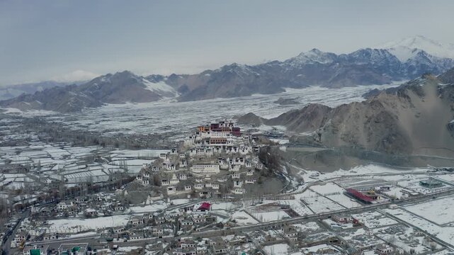 Thiksey Monastery rises above the Indus Valley in winter, surrounded by snow-covered fields and Himalayan peaks. Smooth aerial orbit reveals the Tibetan Buddhist complex and frozen agricultural.