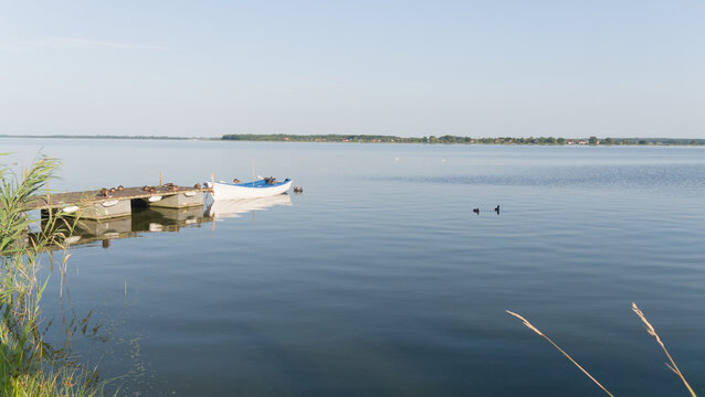 Mała biała ł&oacute;dka przy pomoście na spokojnym jeziorze o poranku, kaczki na wodzie.