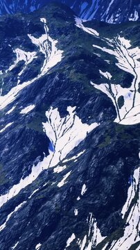 A view of a mountain landscape features rocky slopes, patches of snow, and areas of green vegetation. The scene captures the natural beauty and rugged terrain of the area.