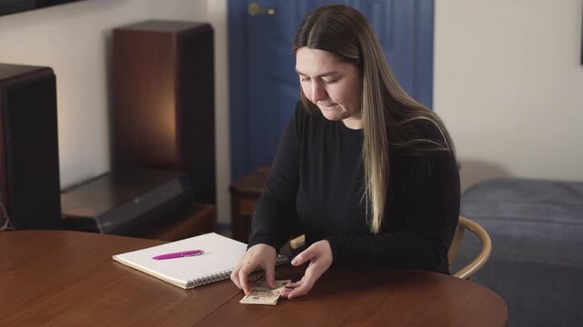 Young woman reviews budget notes and cash while setting up a monthly budget at a home table.