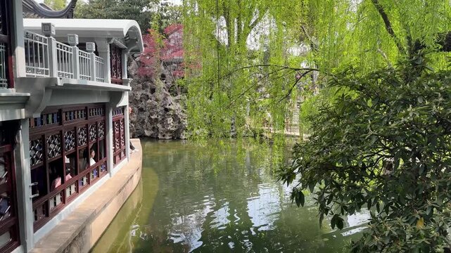 Suzhou, China, 01 April 2026 ? A tranquil pond reflects weeping willows and traditional Chinese pavilion with ornate lattice windows beside Taihu rockery in spring.