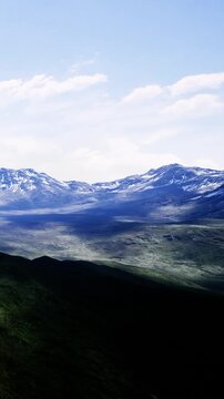 A vast landscape shows mountains under a clear sky. The scene captures the play of light and shadow on the rugged terrain. Snow covers the peaks, creating contrast with green valleys.