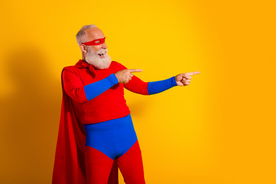 Happy elderly superhero in red and blue costume points with cape against yellow background for fun show promotion