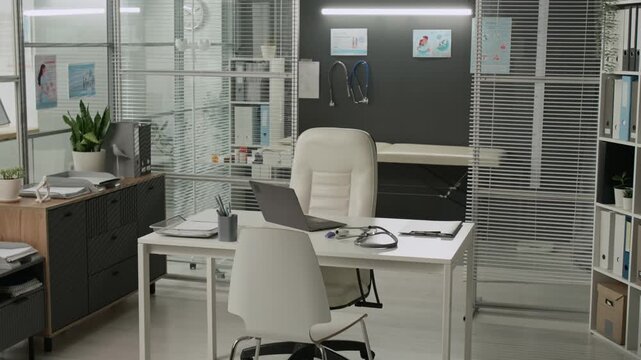 Interior of bright modern doctors office featuring white desk with laptop and stethoscope, two chairs, file storage rack, and medical couch in examination room in background