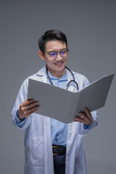 Asian doctor in a white coat with a stethoscope and glasses, using a digital tablet, pointing on the screen with his finger, on a gray background.