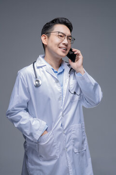 Asian doctor in a white coat with a stethoscope and glasses, isolated on a grey background, talking on a smartphone with a smiling expression and one hand in his pocket.