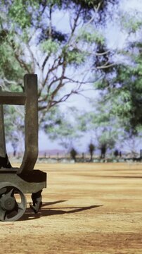 A cart stands on a dirt path surrounded by trees and open space. The sun shines down, and the sky is clear. The scene shows nature and a simple object in a rural area.