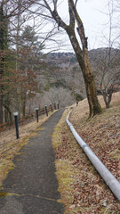 Fototapeta premium Industrial water pipeline running alongside a rural asphalt walkway on a hillside, scenic outdoor infrastructure in a mountainous Japanese village during winter.