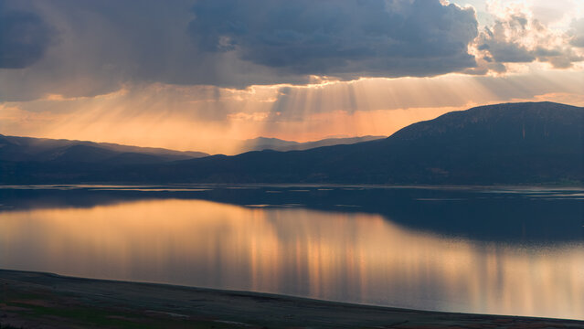 Beams of divine light filtering through the clouds at sunset over Lake Burdur. The golden silhouette of the mountains reflected on the water.