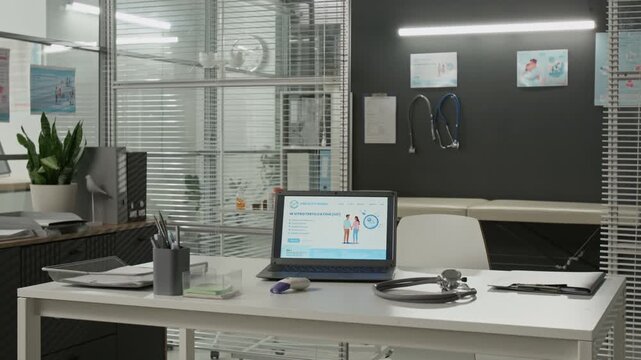 No people view of modern medical office showing white desk with laptop displaying website page of fertility clinic, stethoscope and forehead thermometer, with examination area in background