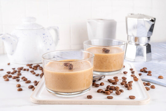 Italian coffee latte mousse dessert, whipped semifreddo souffle or pudding in portioned glass jars, with coffee beans, on white marble table copy space