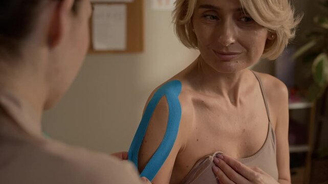Over shoulder closeup of massage therapist applying blue kinesiology tape on female client shoulder, showing muscle support and rehabilitation treatment in wellness clinic