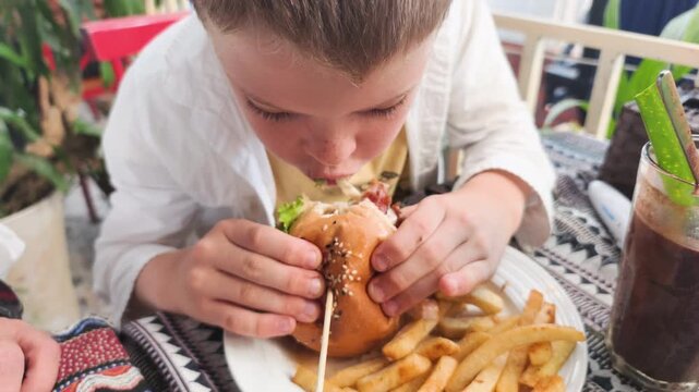 Biting delicious burger shows young boy enjoying big bite of juicy cheeseburger. French fries and cold drink sit on patterned tablecloth in outdoor cafe. This biting delicious burger moment.