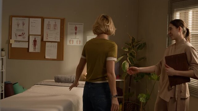 Medium shot of Caucasian woman entering cozy treatment room and being welcomed by female massage therapist, having brief conversation before starting therapeutic session in calm wellness setting