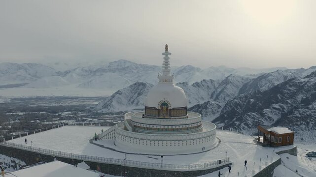 Shanti Stupa stands on a snow-covered hilltop surrounded by Himalayan mountain peaks in Leh, Ladakh. Smooth orbital camera movement circles the white Buddhist monument revealing the winter landscape.