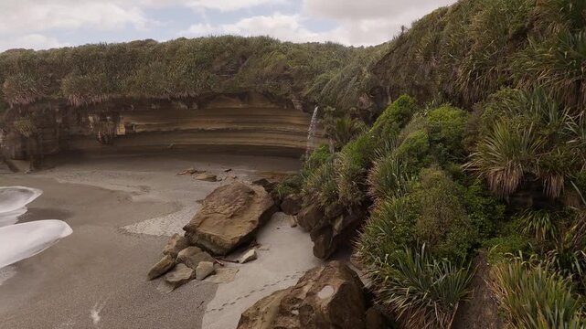 Waterfall flows from lush green cliff face directly onto sand of Punakaiki remote beach on Truman Track, New Zealand. Aerial drone
