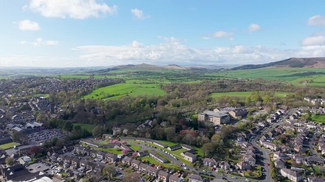 A reversing drone shot pulls back from the Skipton Building Society headquarters, revealing northern Skipton's residential streets, green fields and rolling moorland under blue skies. Spring.