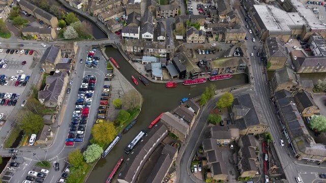A drone tilts down over Skipton's historic canal basin on the Leeds and Liverpool Canal, revealing vivid red leisure cruise boats, stone buildings, car parks and converging roads. North Yorkshire, spr