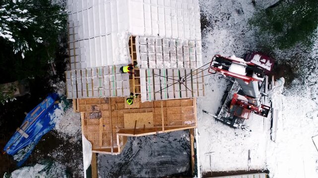 Top down drone view of roof construction with workers installing structure panels using crane. Winter building site showing timber framework and residential development process.