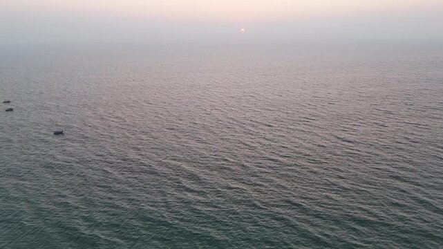 Cinematic aerial view of a vast open sea under a hazy sunset, where gentle ripples stretch endlessly and the fading sun appears as a soft glow near the horizon, creating a minimal and tranquil.