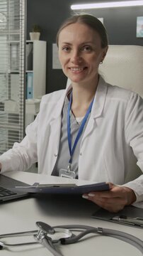 Vertical portrait of female physician looking at camera with smile while analyzing medical records sitting at white table